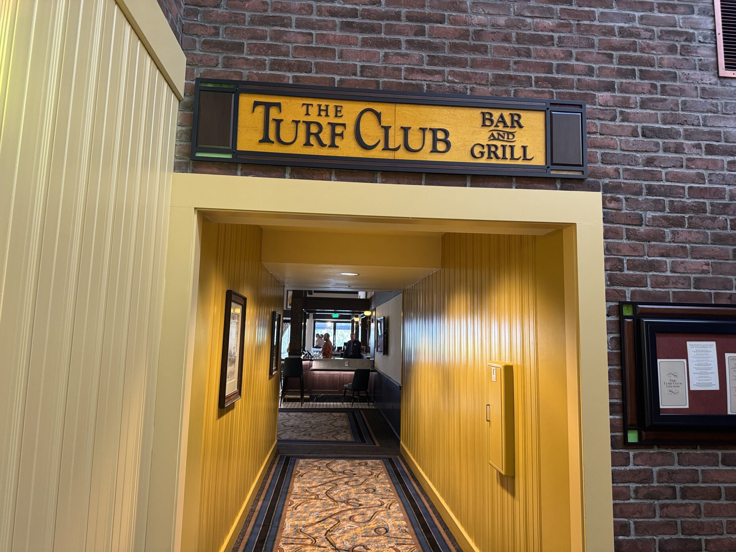 A carved yellow sign reading The Turf Club Bar and Grill mounted on red brick above a yellow paneled corridor leading into the lounge