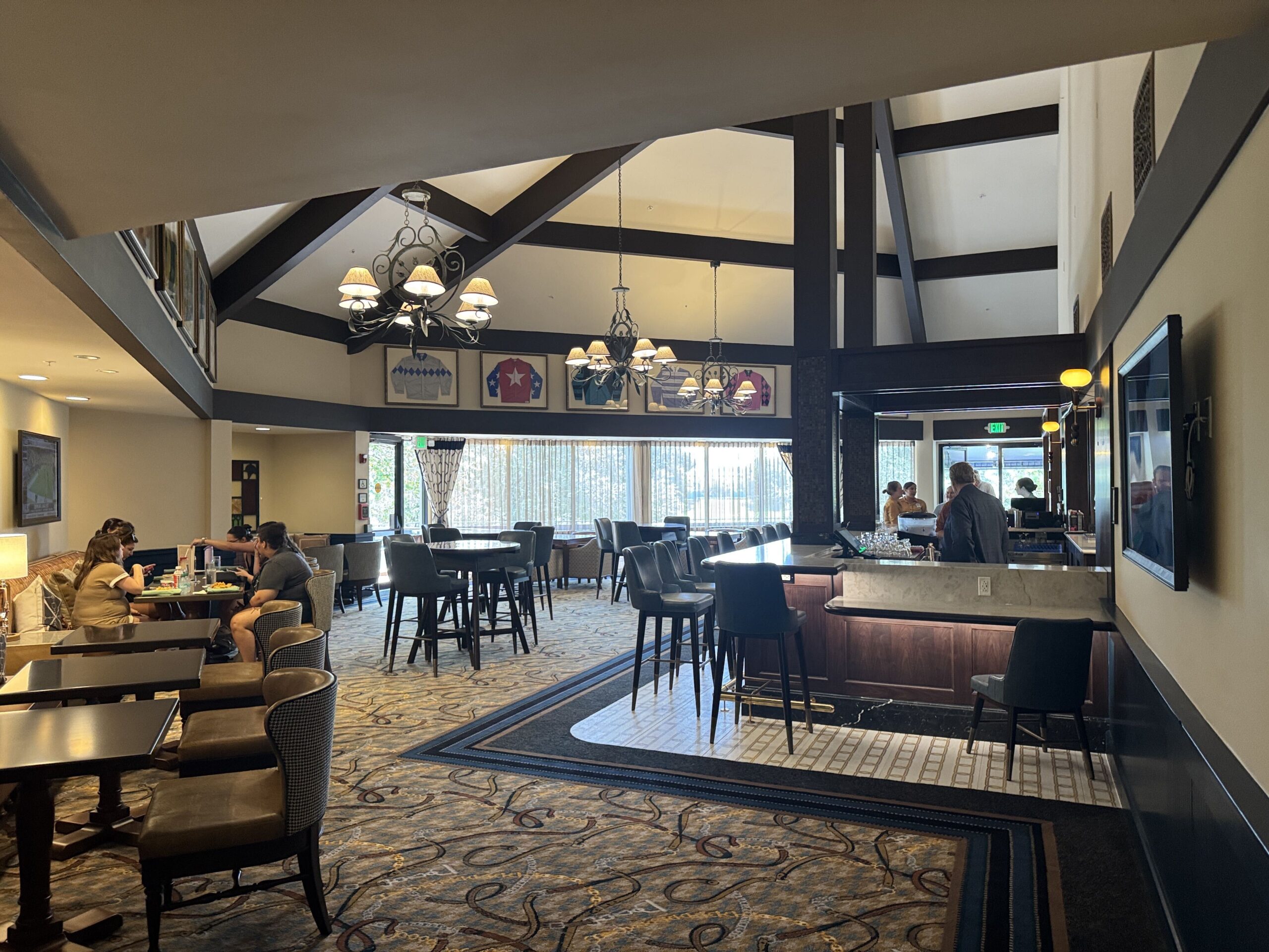 The reopened lounge interior with a full bar built out along the right side, faux leather barstools, a vaulted cream ceiling with dark beams, three iron chandeliers and framed jockey silks on the wall