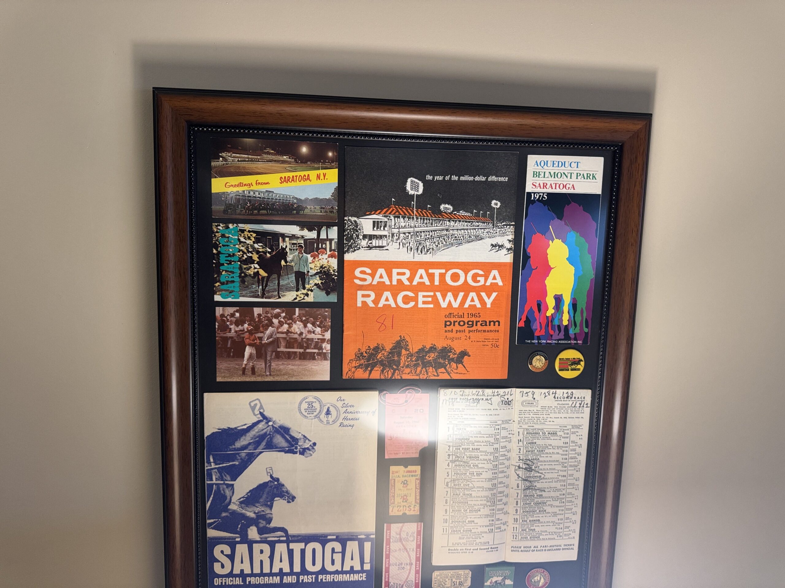 A dark wood frame filled with Saratoga racing memorabilia including a 1965 Saratoga Raceway program, a 1975 New York Racing Association program, postcards, Saratoga Harness pins and old raceway ticket stubs