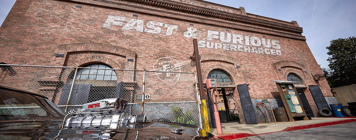 The brick warehouse facade of Fast & Furious: Supercharged with the ride name painted in large white letters, a chain link fence in front, a virtual line kiosk and a ride entrance doorway, with a chrome engine supercharger on a black car hood in the foreground