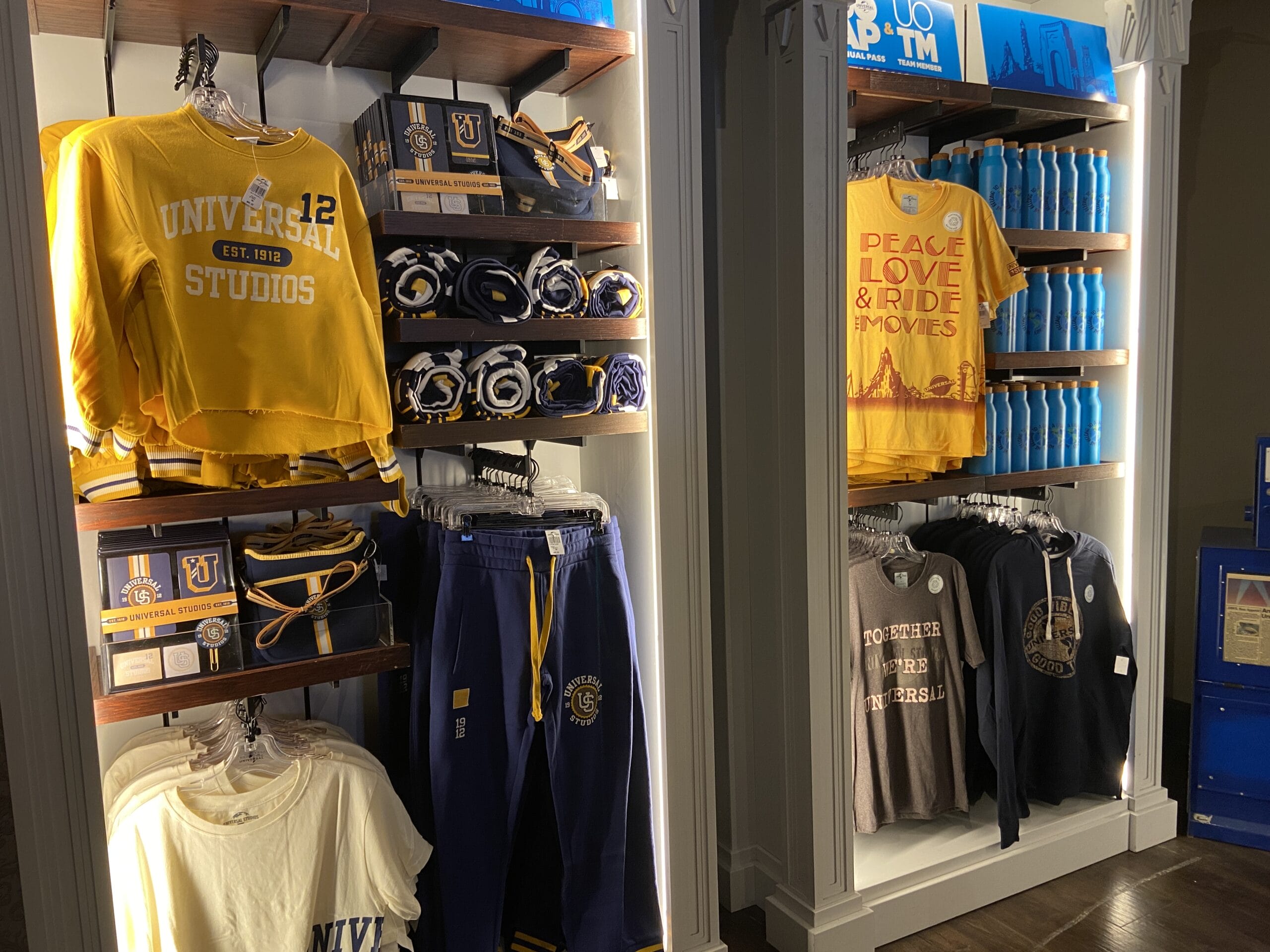 Shop shelving holding a yellow Universal Studios Est. 1912 sweatshirt, rolled blankets, navy sweatpants, folded tee shirts, a Peace Love and Ride the Movies shirt and a row of blue water bottles, with a blue UOAP and UOTM sign on the top shelf