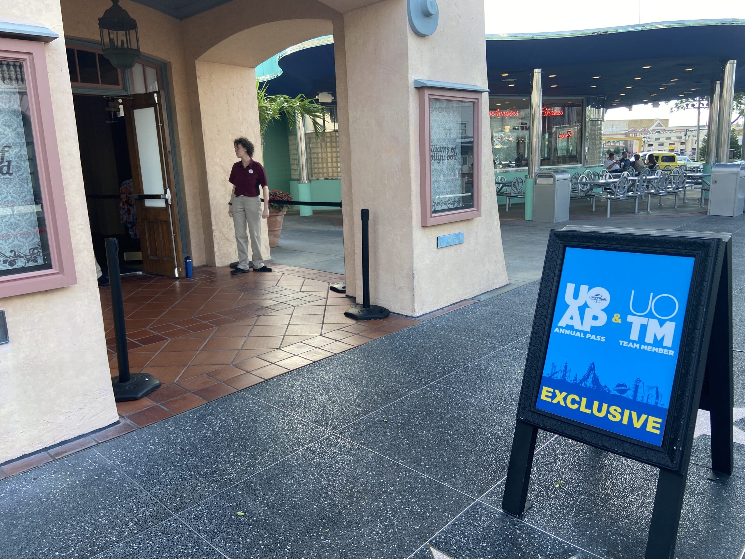 A blue A-frame sign in a black frame on a park walkway reading UOAP Annual Pass and UOTM Team Member above the word EXCLUSIVE in yellow, with a Universal Orlando Resort logo, standing outside an open shop doorway