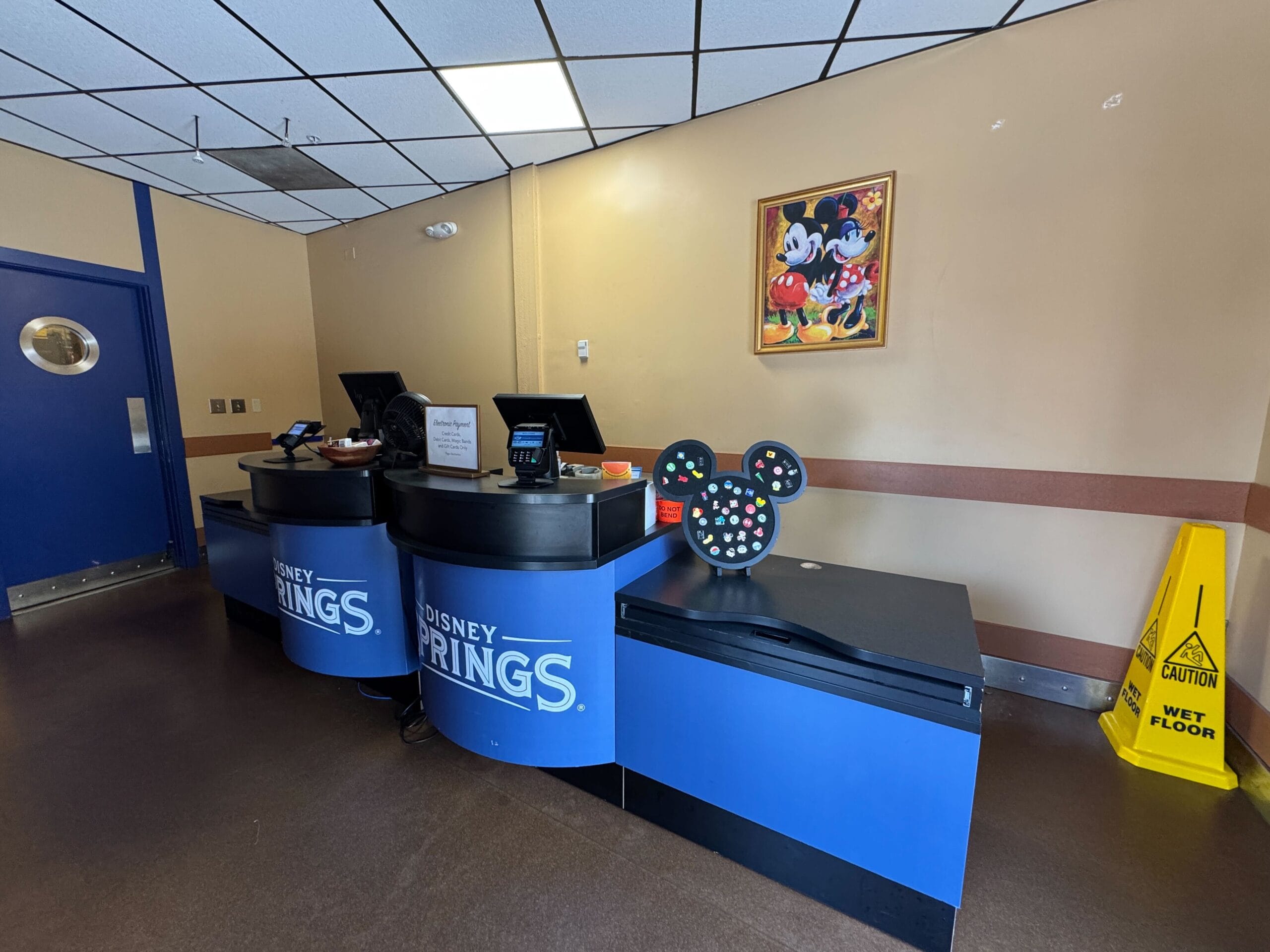 A back room counter with two blue Disney Springs registers, payment terminals, a Mickey-shaped pin display board and a framed Mickey and Minnie painting on a tan wall