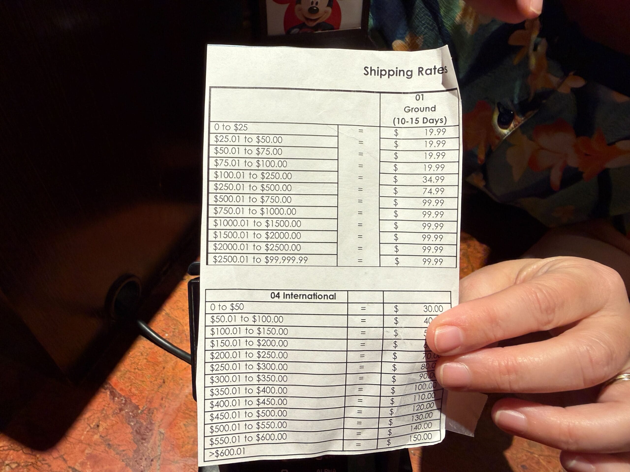 A hand holding a paper Shipping Rates card showing value bands from 0 to $25 through $2500.01 to $99,999.99 against a Ground 10-15 Days price column