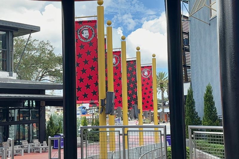 Tall star banners hanging from yellow poles along a CityWalk walkway, most of them red with black stars and carrying the circular Fat One's logo, with one reversed to black with red stars, under a blue sky with palm trees behind.