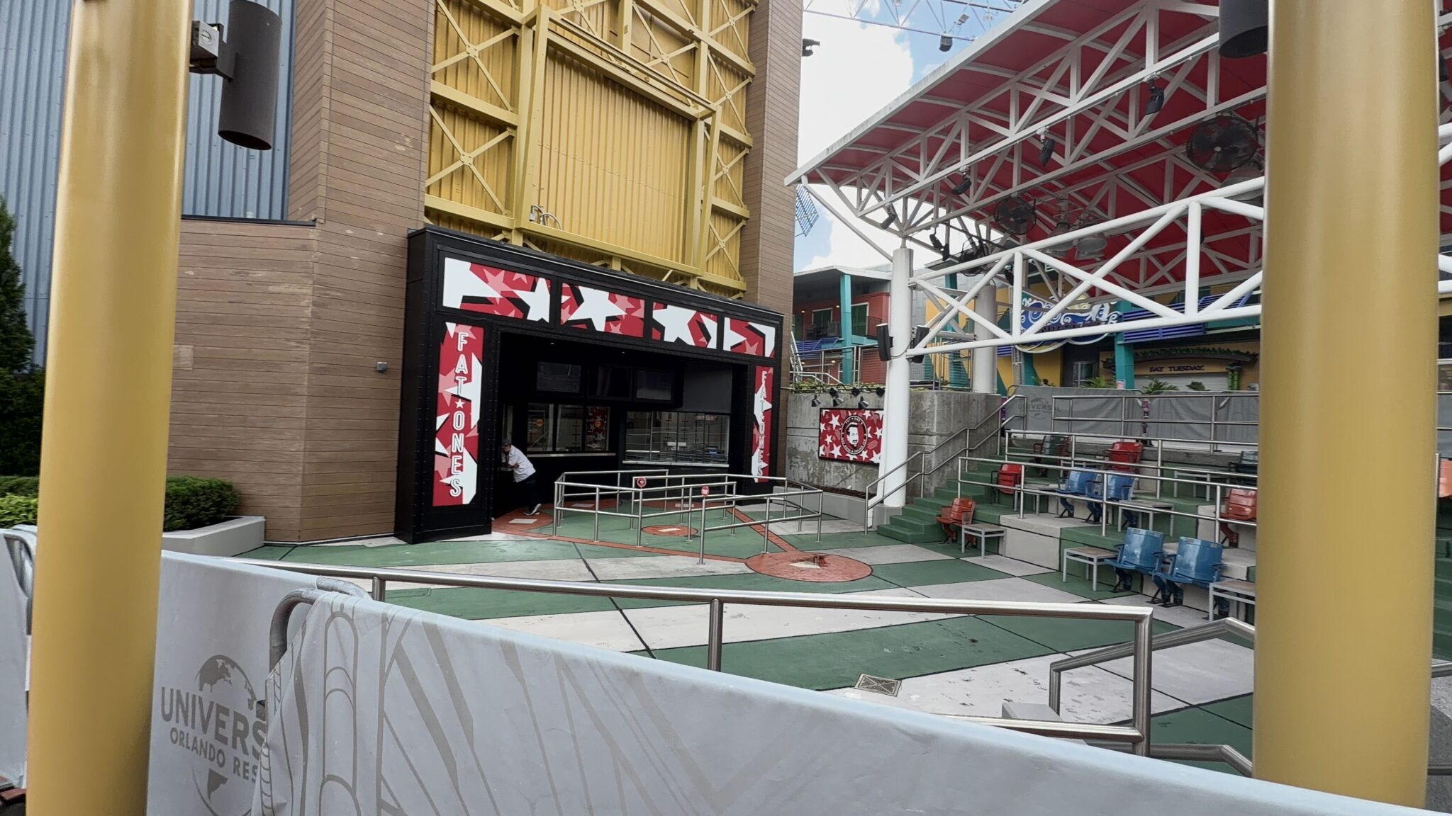 The Fat One's storefront at Universal CityWalk seen past a white Universal Orlando Resort construction barricade, with white vertical FAT ONE'S lettering on red star-patterned panels flanking a black framed entrance and red, white and black star graphics across the header, beside the former stadium seating with red and blue ballpark seats on tiered risers.