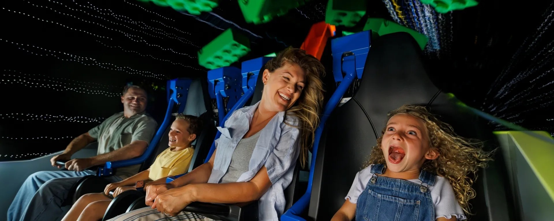 A family riding Galacticoaster inside a darkened coaster building, a woman laughing with her hair blown back and a girl in denim overalls screaming with delight