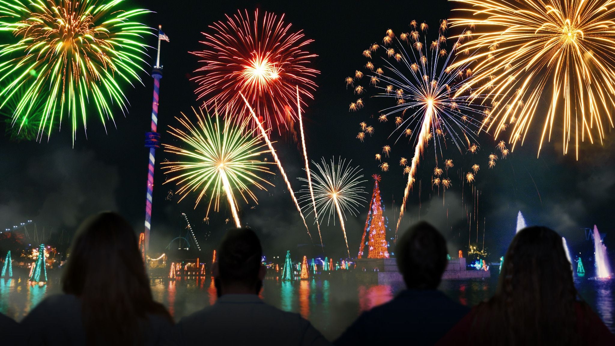 Four guests silhouetted watching green, red and gold fireworks burst over a lagoon with a large lit Christmas tree on an island