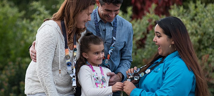 Two adults and a young girl wearing lanyards covered in collectible pins stand talking with a SeaWorld team member in a teal jacket, who is holding out her own pin covered lanyard, with green and red foliage behind them.