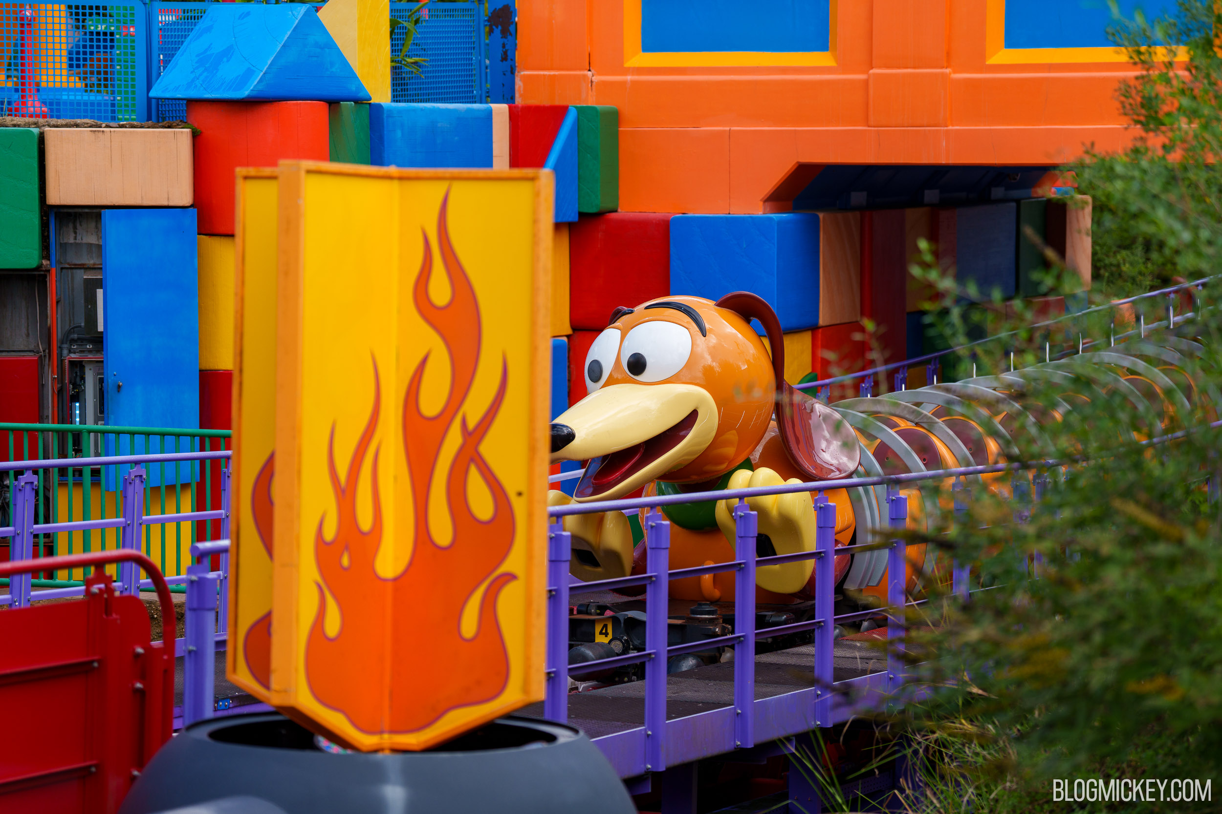 A Slinky Dog Dash ride vehicle stopped on the track behind purple railings, with a yellow flame-painted scenery flat in the foreground and an open blue access door in the block wall at far left.