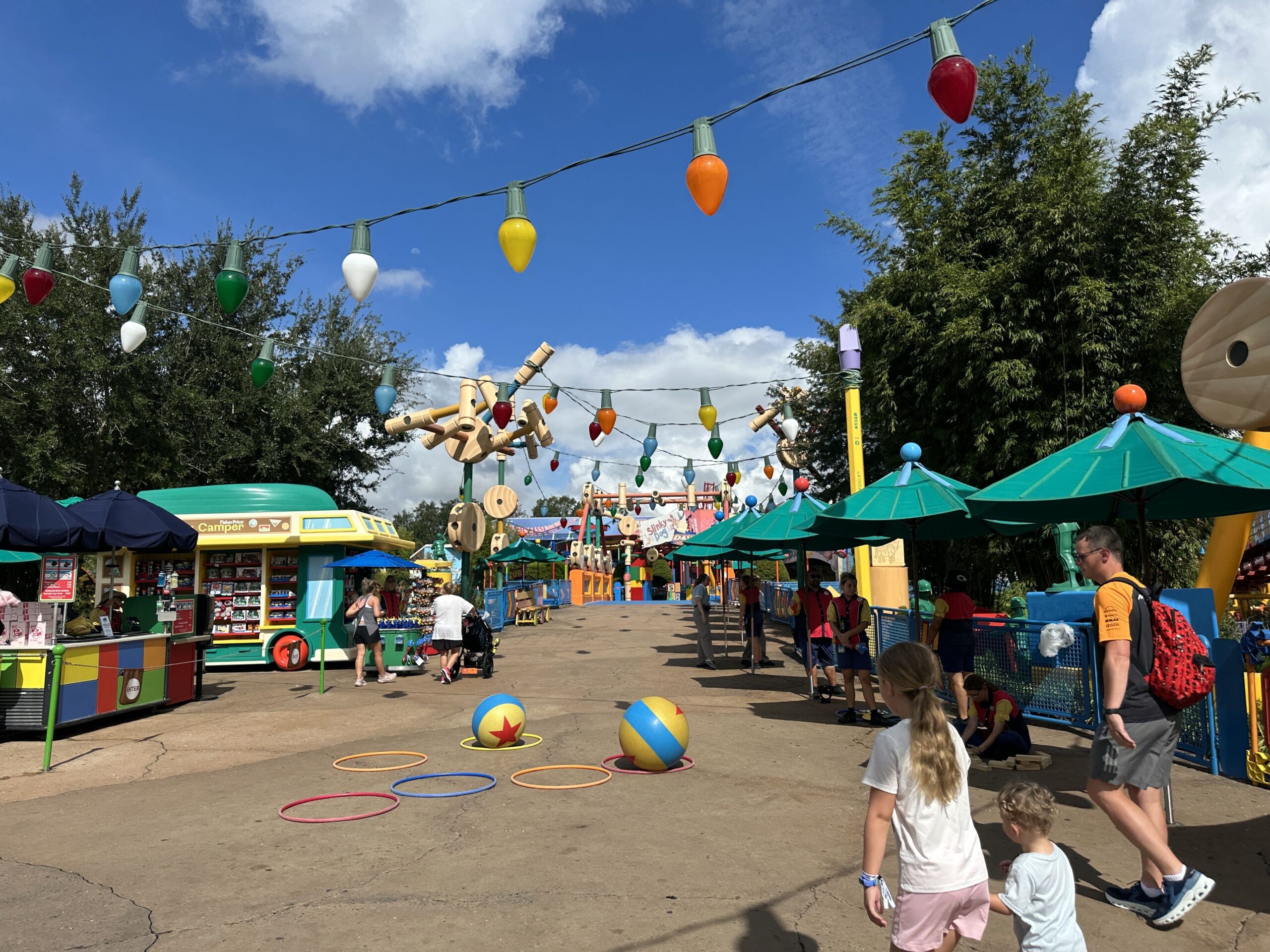 Two giant yellow and blue Pixar balls sitting inside colored hula hoops laid out on the pavement in Toy Story Land, with Cast Members in red and yellow costumes standing along the railing at right and oversized string lights overhead.