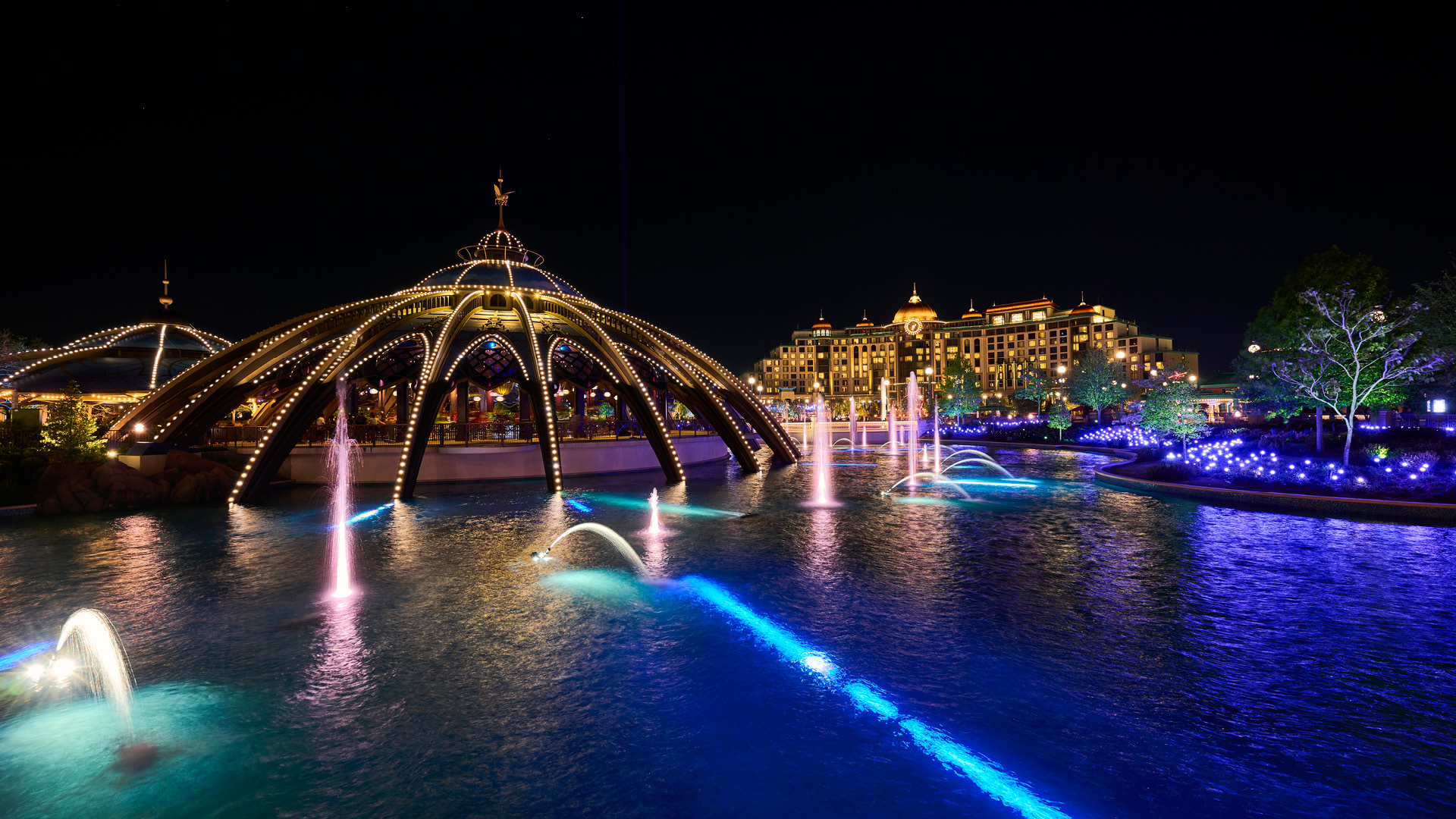 Illuminated fountains in pink, teal and white arcing over the lit blue water of the Celestial Park lagoon at night