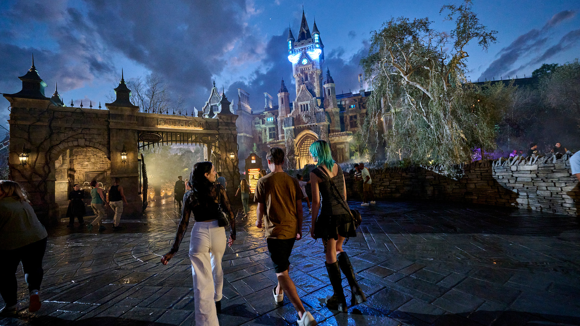 Guests walking toward the Frankenstein gate in Dark Universe, the manor spires lit in blue with fog rolling out through the archway