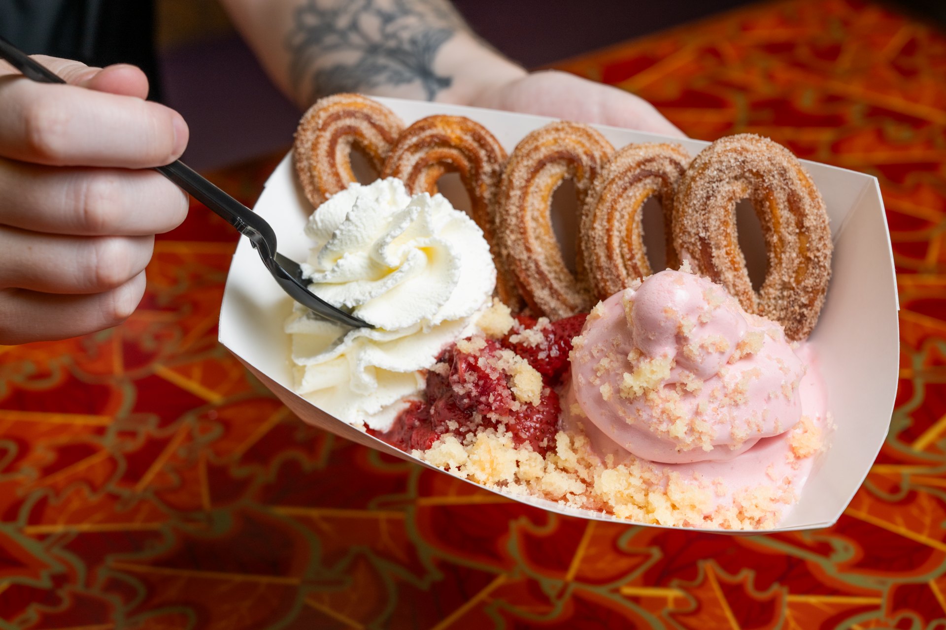 A paper boat holds churro loops, pink strawberry soft serve, strawberry compote, whipped cream and cake crumble, held over a red patterned table.