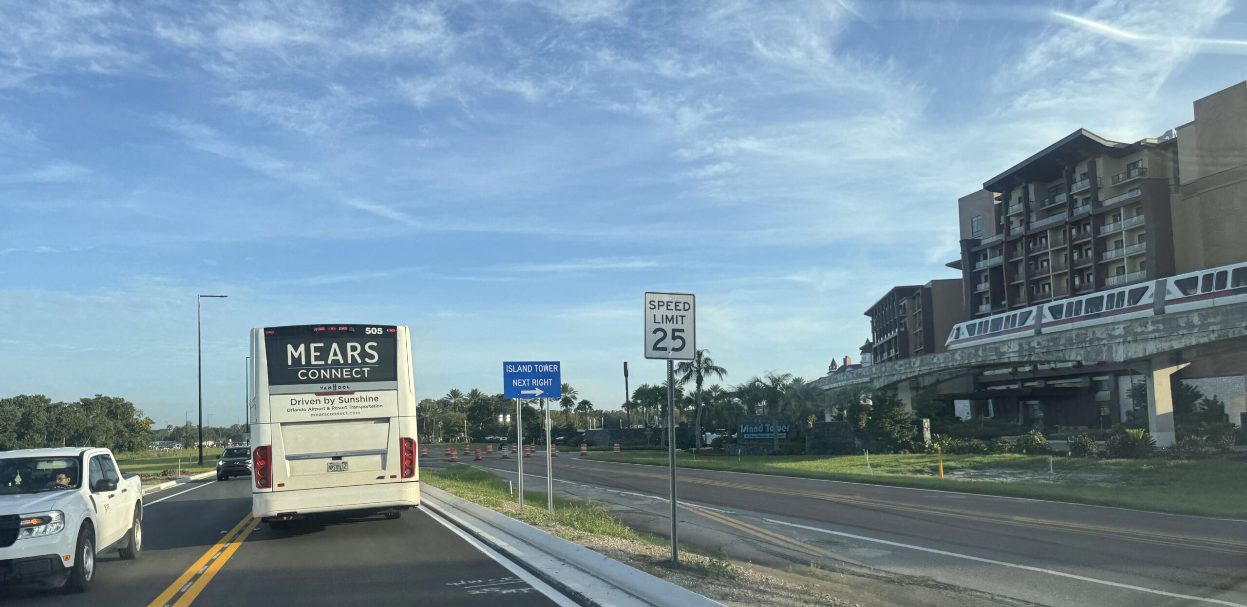 Driving south on the new Floridian Way behind a Mears Connect motorcoach, past a blue sign reading ISLAND TOWER NEXT RIGHT and a 25 mile per hour speed limit sign, with the Island Tower at Disney's Polynesian Village Resort and a monorail on the beam at right.