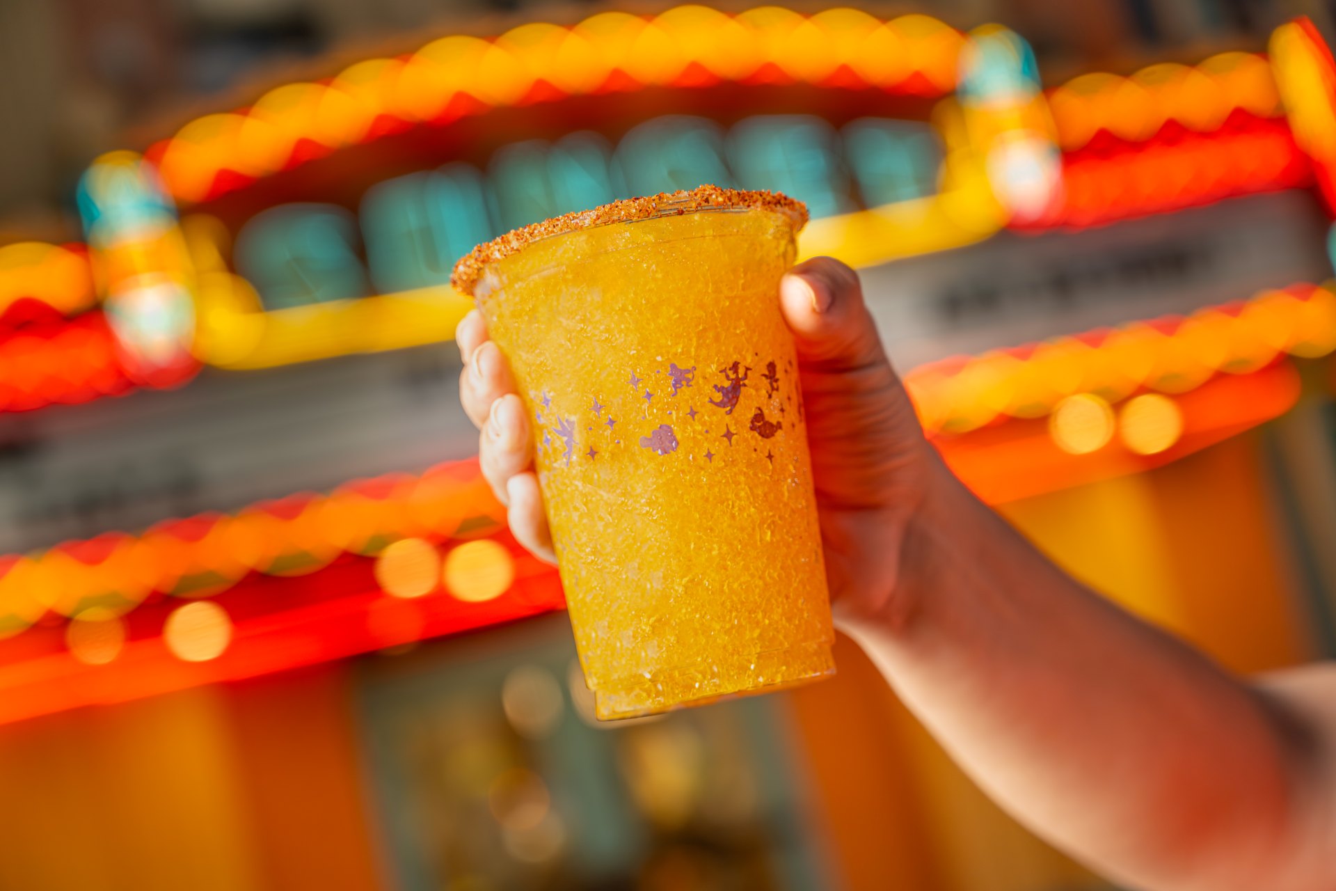 A hand holds a tall clear cup of golden orange frozen slush with a chili and salt rim, printed with small purple characters and stars, in front of a blurred neon marquee.