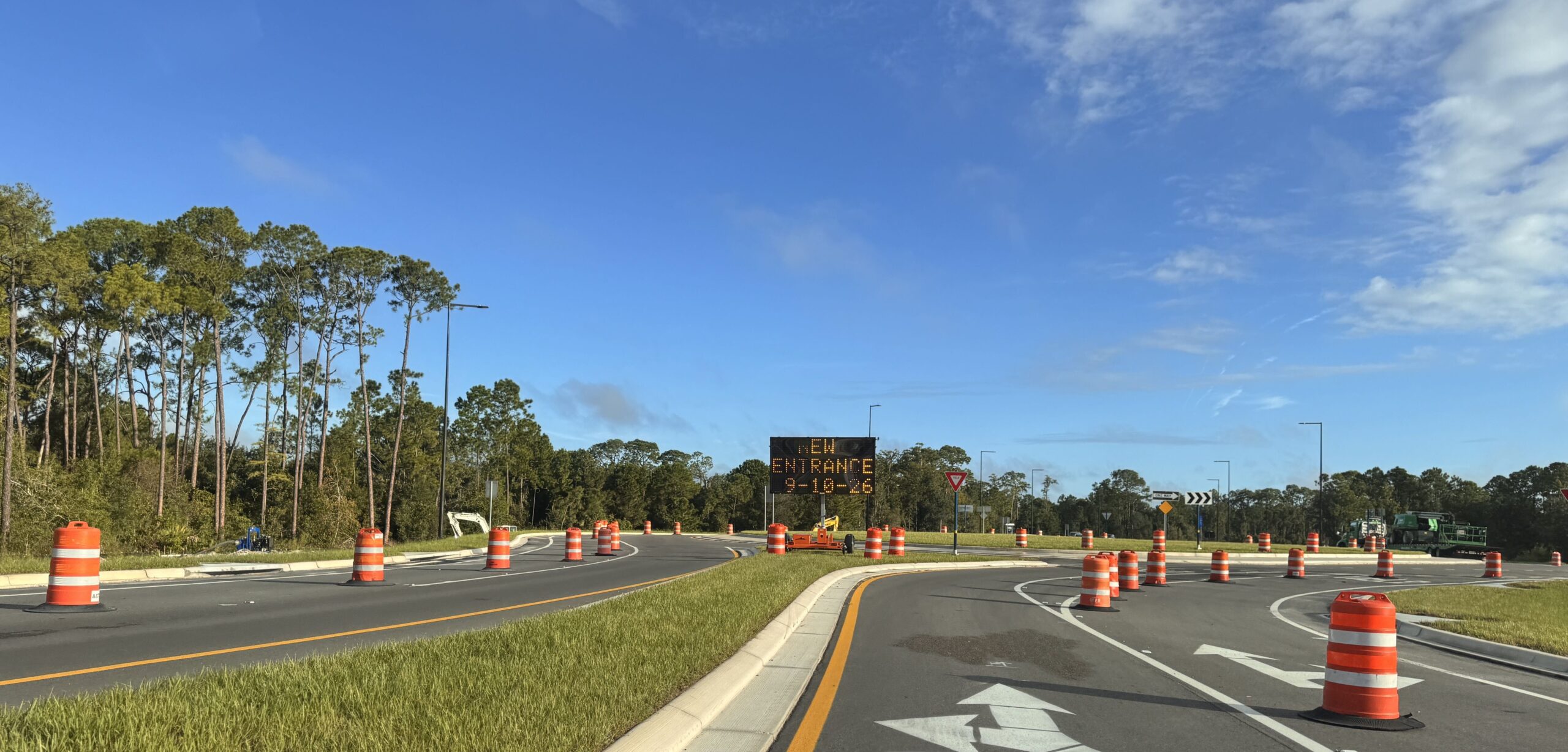 An amber LED construction message board reading NEW ENTRANCE 9-10-26 stands beside a new roundabout on Floridian Way, surrounded by orange barrels, with an excavator parked at left.