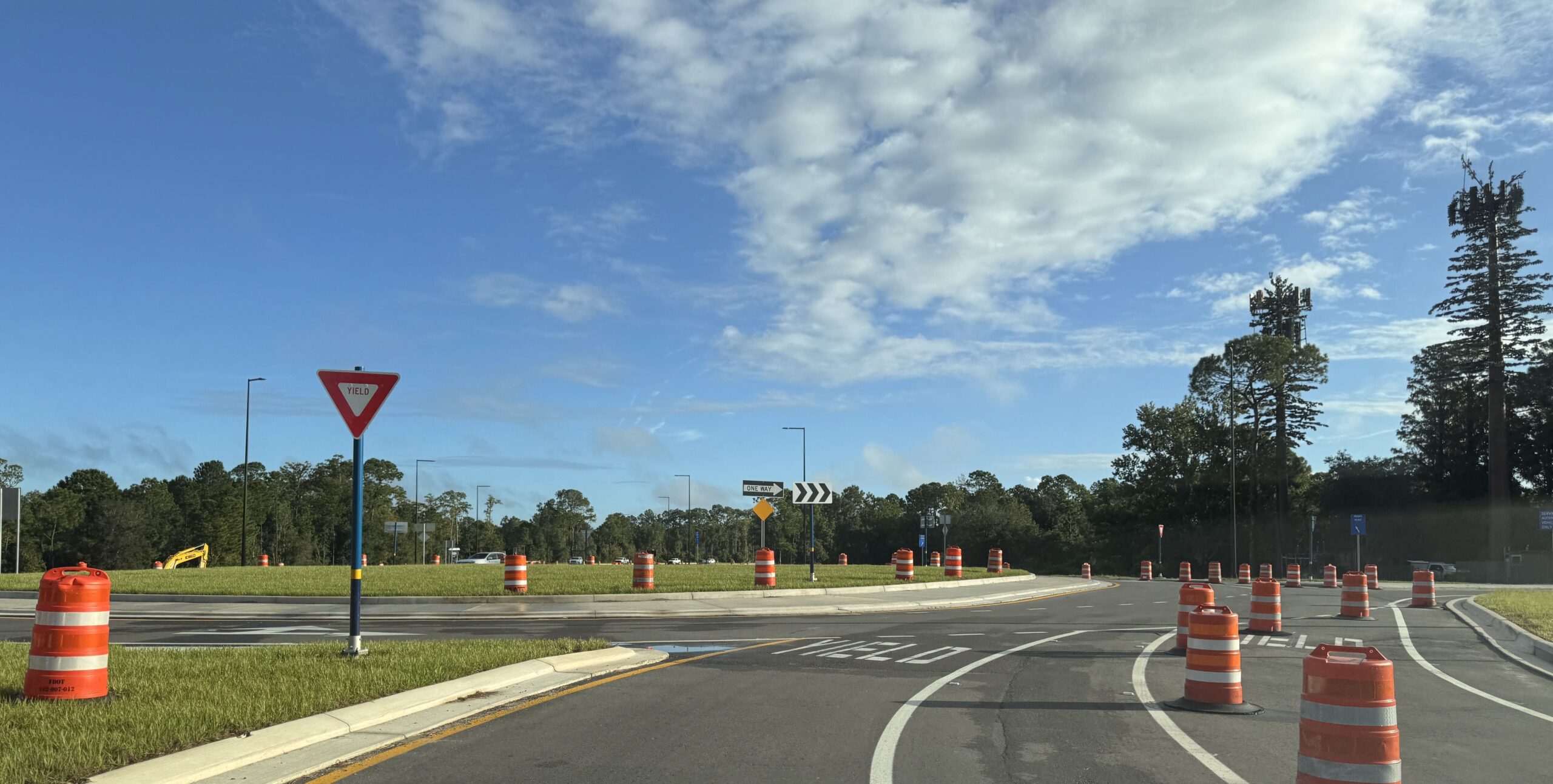 A ground level view of the new roundabout on Floridian Way, with a yield sign on a blue pole, one way and chevron signs on the far side, orange barrels lining the splitter islands, and a cell tower disguised as a pine tree at right.
