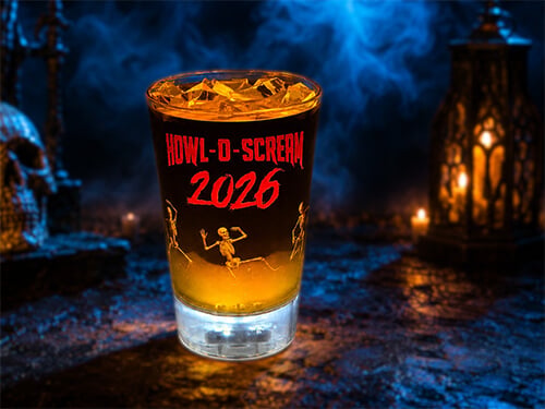 An amber cocktail over ice in a glowing light-up souvenir cup printed with HOWL-O-SCREAM 2026 in red lettering and dancing skeletons, photographed on a foggy graveyard set with a skull at left and a lit lantern at right.