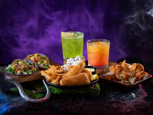 Howl-O-Scream food and drink spread on a purple smoke filled background: two tacos on a dark plate, a black plate of battered fried fish and french fries, a square wooden dish of seasoned shrimp topped with shaved cheese and herbs, a green cocktail and an orange cocktail.