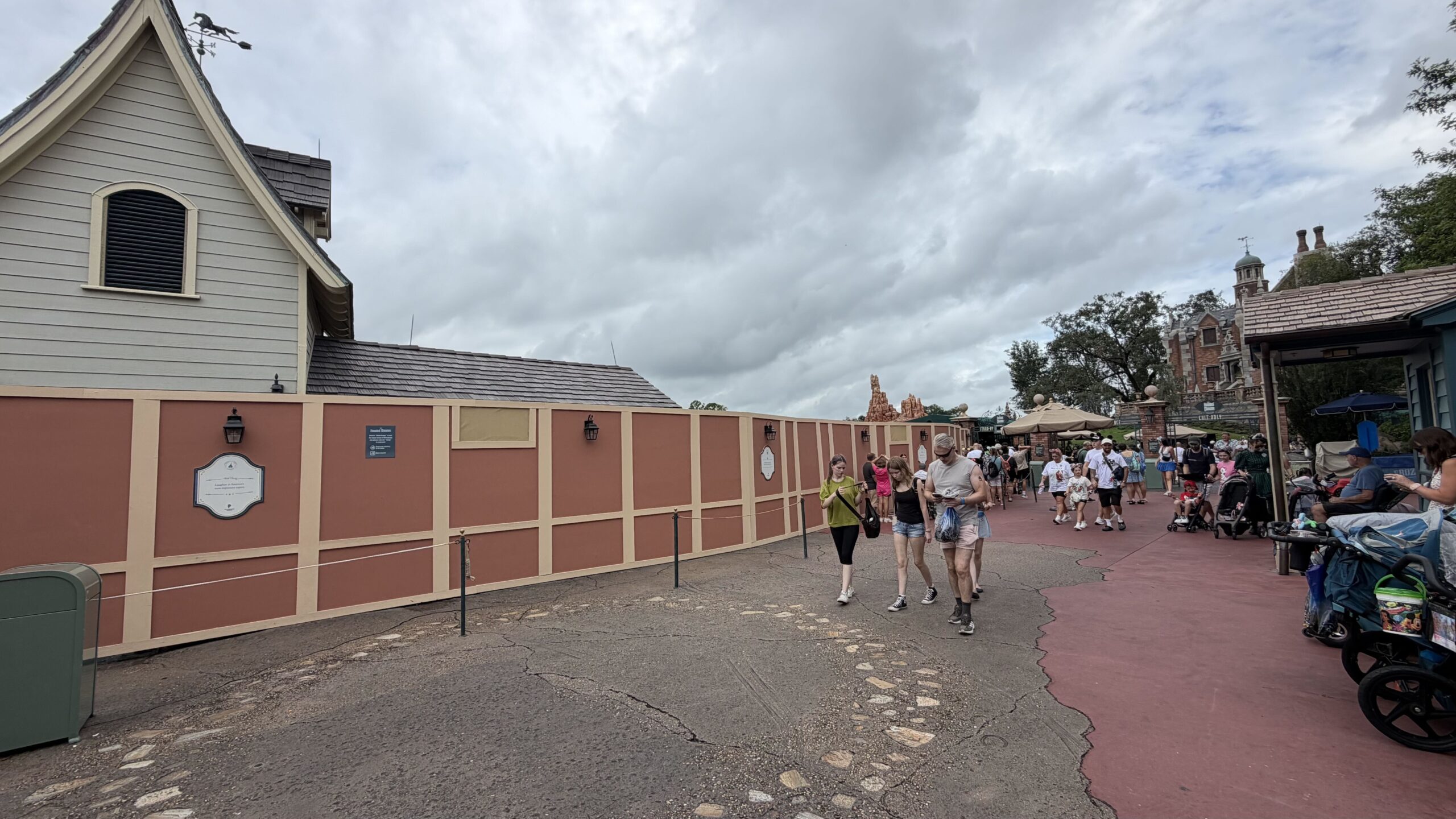 A long rust colored construction wall with cream trim running past a grey gabled building, with the red rock peaks of Big Thunder Mountain Railroad visible in the distance and the Haunted Mansion facade at right