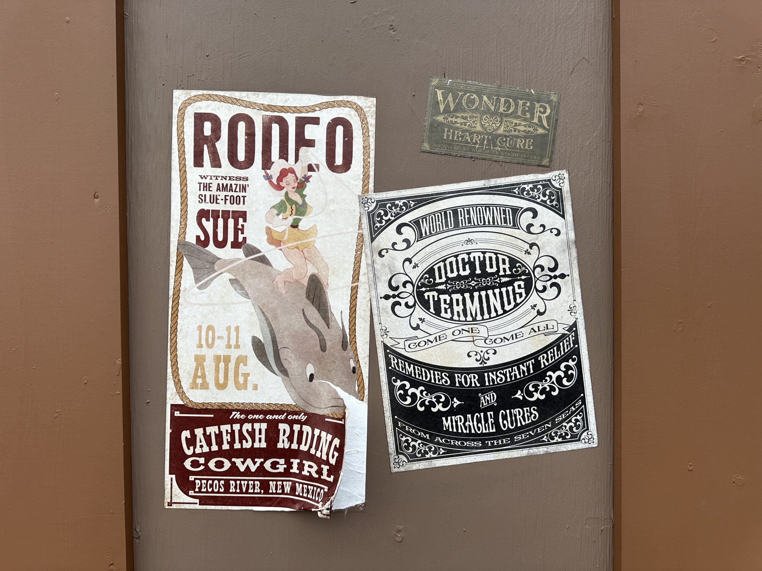 Two paper posters on a brown wall, one a rodeo bill for The Amazin Slue-Foot Sue showing a cowgirl riding a catfish, the other a black and white handbill for Doctor Terminus remedies, with a small metal Wonder Heart Cure sign above