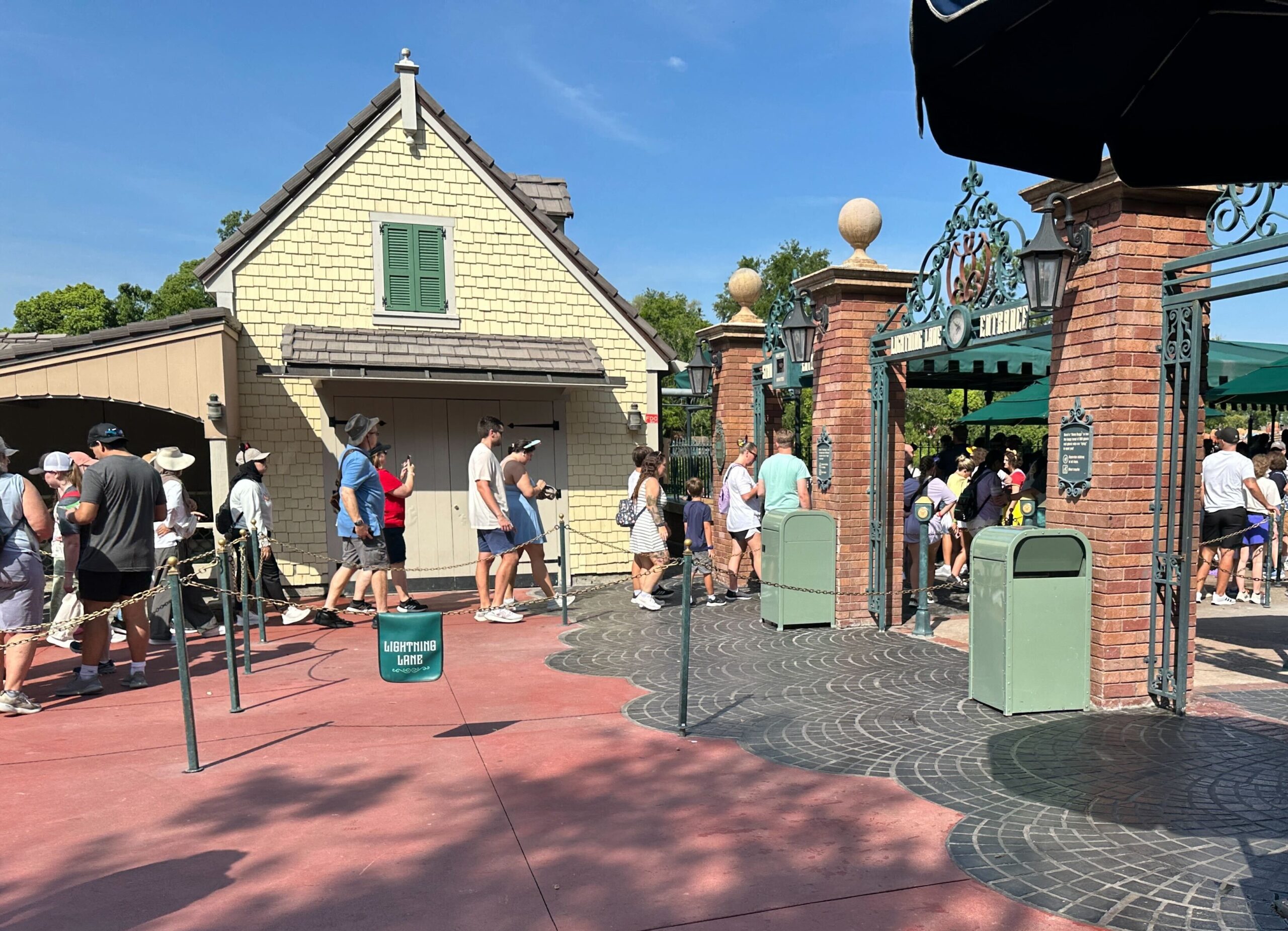 A pale yellow shingled building with a single green louvered shutter and a shed roof over closed double doors, with guests walking past on a red pathway and an ornate green metal arch marked Lightning Lane Entrance at right