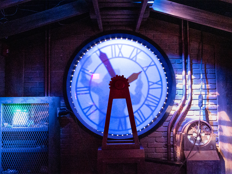 A large backlit clock face glowing blue behind a red gear topped frame, with Doc Brown's silhouette visible against the dial, set into a brick wall hung with conduit and machinery.