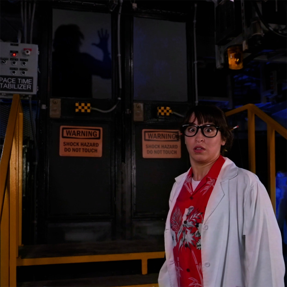 A wide eyed woman in a white lab coat over a red Hawaiian print shirt with black framed glasses and a pencil behind her ear, standing beside doors marked WARNING SHOCK HAZARD DO NOT TOUCH, with a silhouetted figure pressing a hand against the glass behind her.