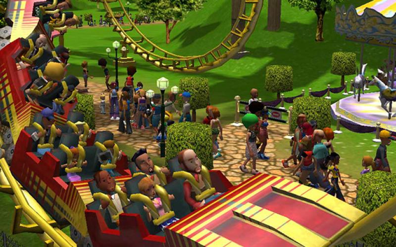 RollerCoaster Tycoon 3 gameplay showing a red and yellow coaster train cresting a drop full of riders, with park guests on winding paths, trimmed hedges and a carousel.
