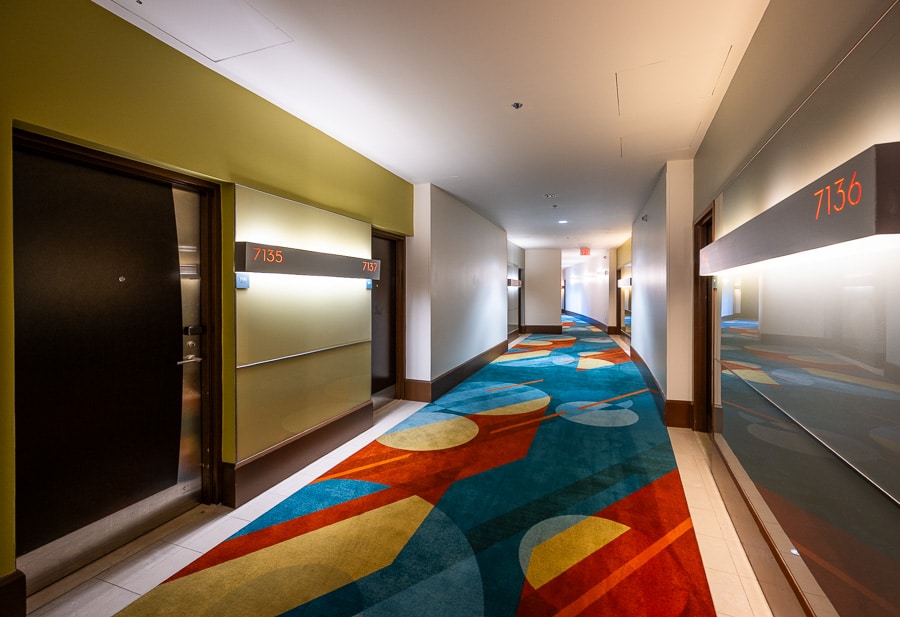 A hotel corridor with a boldly patterned blue, red and yellow carpet, olive and grey walls, and backlit grey room number panels reading 7135, 7137 and 7136 beside dark guest room doors
