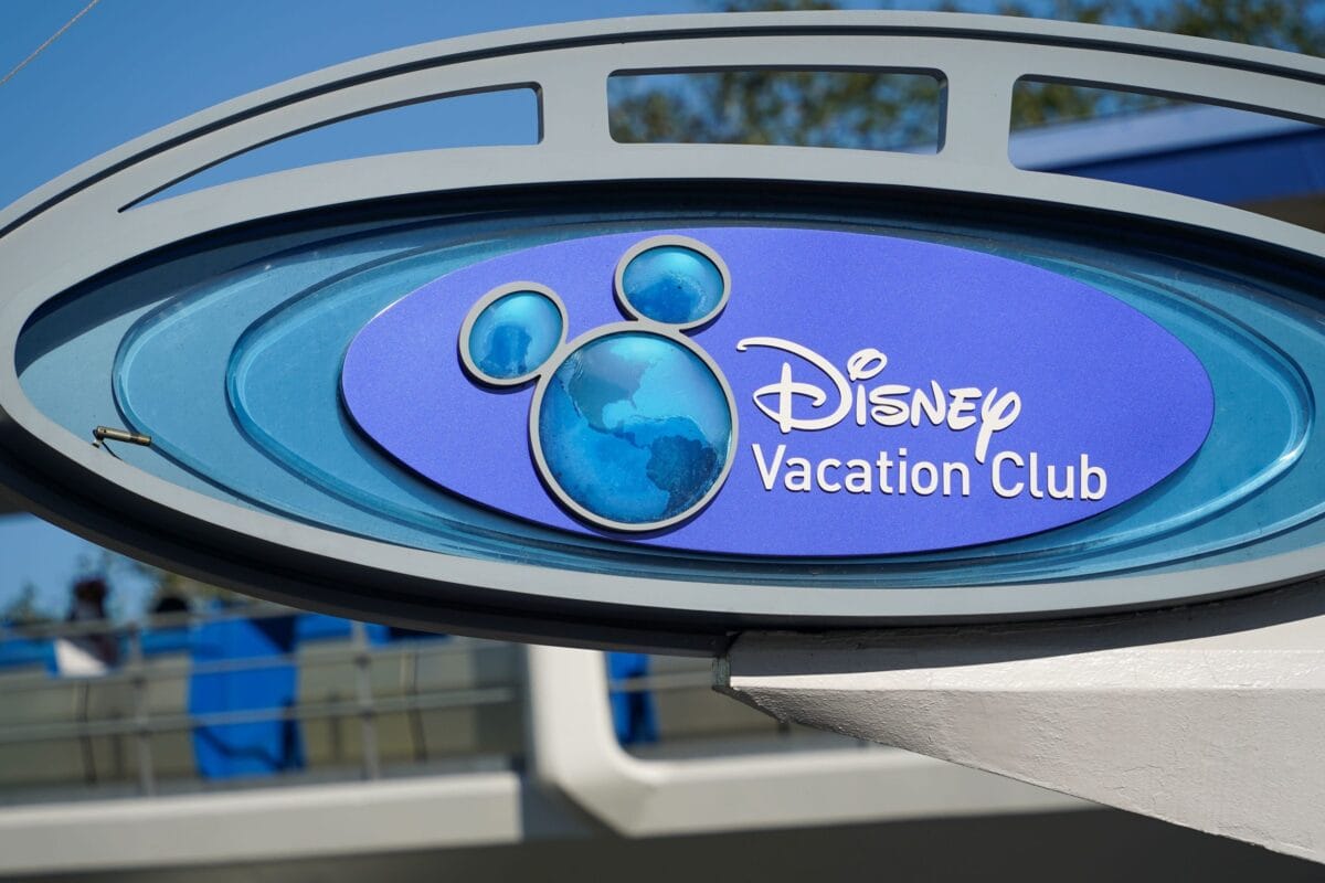 A blue oval Disney Vacation Club sign mounted in a curved grey metal frame against a clear blue sky, with the pale blue Mickey shaped globe logo at the left and the Disney Vacation Club wordmark in white beside it