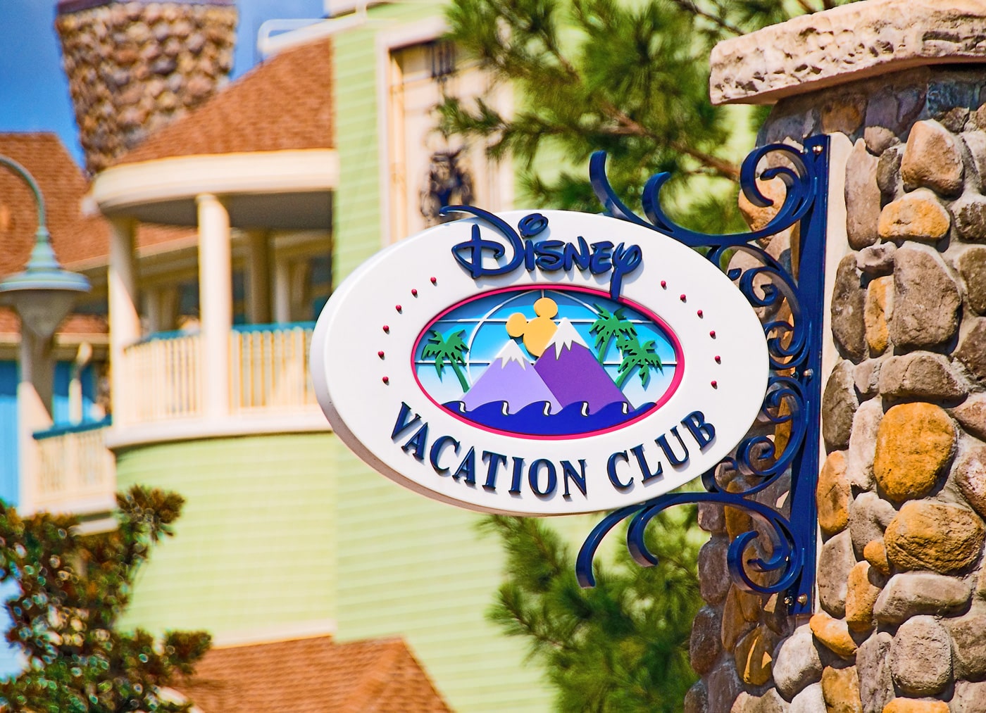 A white oval Disney Vacation Club sign with a crest of purple mountains and green palms on a blue field, hanging from an ornate dark blue metal bracket on a stone pillar, with a green and cream resort building behind it