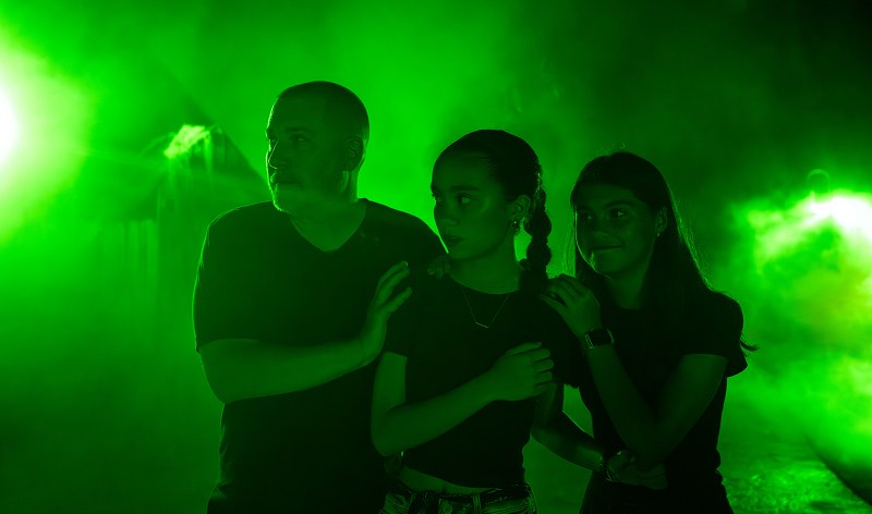 Three guests, a man and two young women, standing close together and looking off camera in heavy green fog lit from behind at Howl-O-Scream