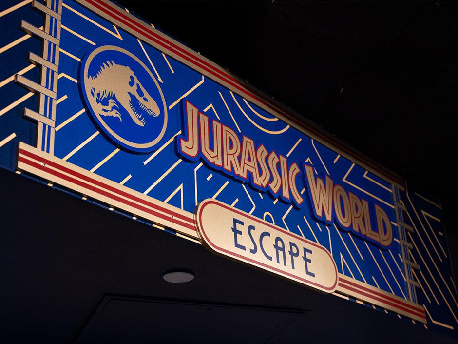 An illuminated blue and gold marquee sign photographed from below, showing a tan dinosaur skull inside a circle beside the words Jurassic World, with the word Escape on an oval plaque beneath