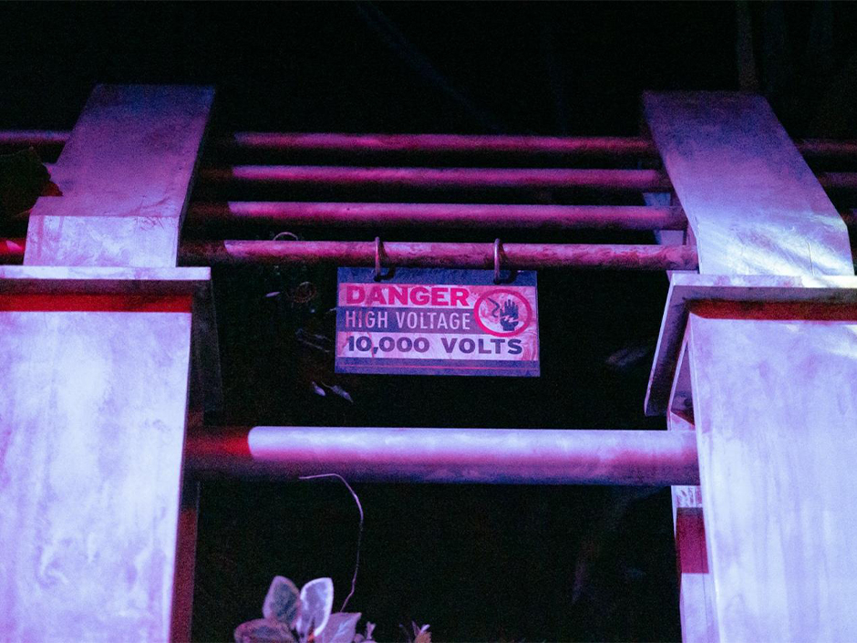 A paddock fence with weathered pillars and metal crossbars lit in deep magenta against a dark background, with a sign hanging from the crossbar reading Danger High Voltage 10,000 Volts beside a red circle enclosing a dark hand struck by a lightning bolt