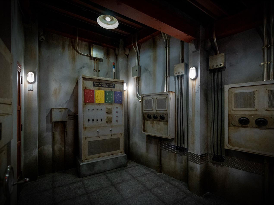 A dim, weathered concrete utility room with exposed conduit and steel junction boxes on the walls, centered on a rusted control cabinet labeled Power Bypass Section C Master Switch Panel with red, yellow, green and blue placards and a row of switches