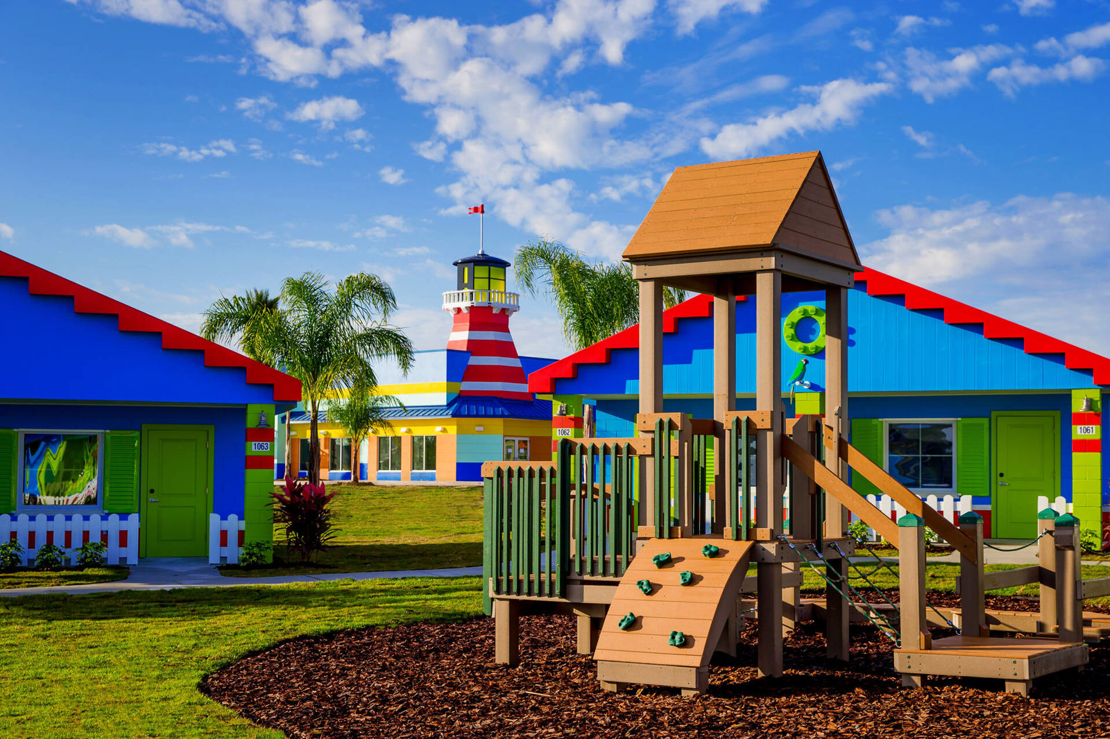 Brightly colored bungalows with red zigzag rooflines and green doors around a lawn, with a wooden play structure in the foreground and a red and white striped lighthouse behind them under a blue sky