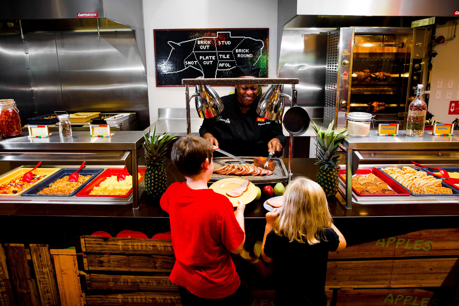 Two children holding plates at a hotel breakfast buffet counter as a chef in a black jacket and cap slices ham under heat lamps, with trays of scrambled eggs, hash browns, pancakes and waffles and a LEGO brick mosaic of a pig on the wall behind