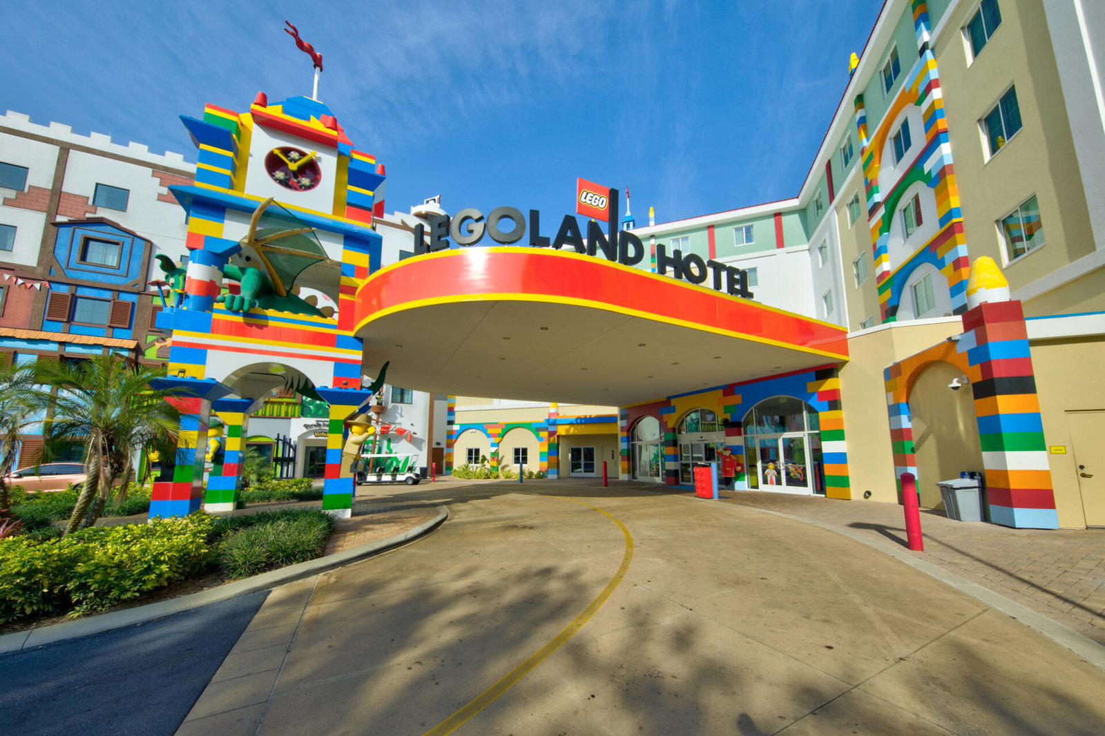 The LEGOLAND Hotel entrance under a bright orange porte cochere, with oversized LEGO brick columns in red, blue, yellow and green, a clock tower topped by a red flag and a large green LEGO dragon mounted on its face