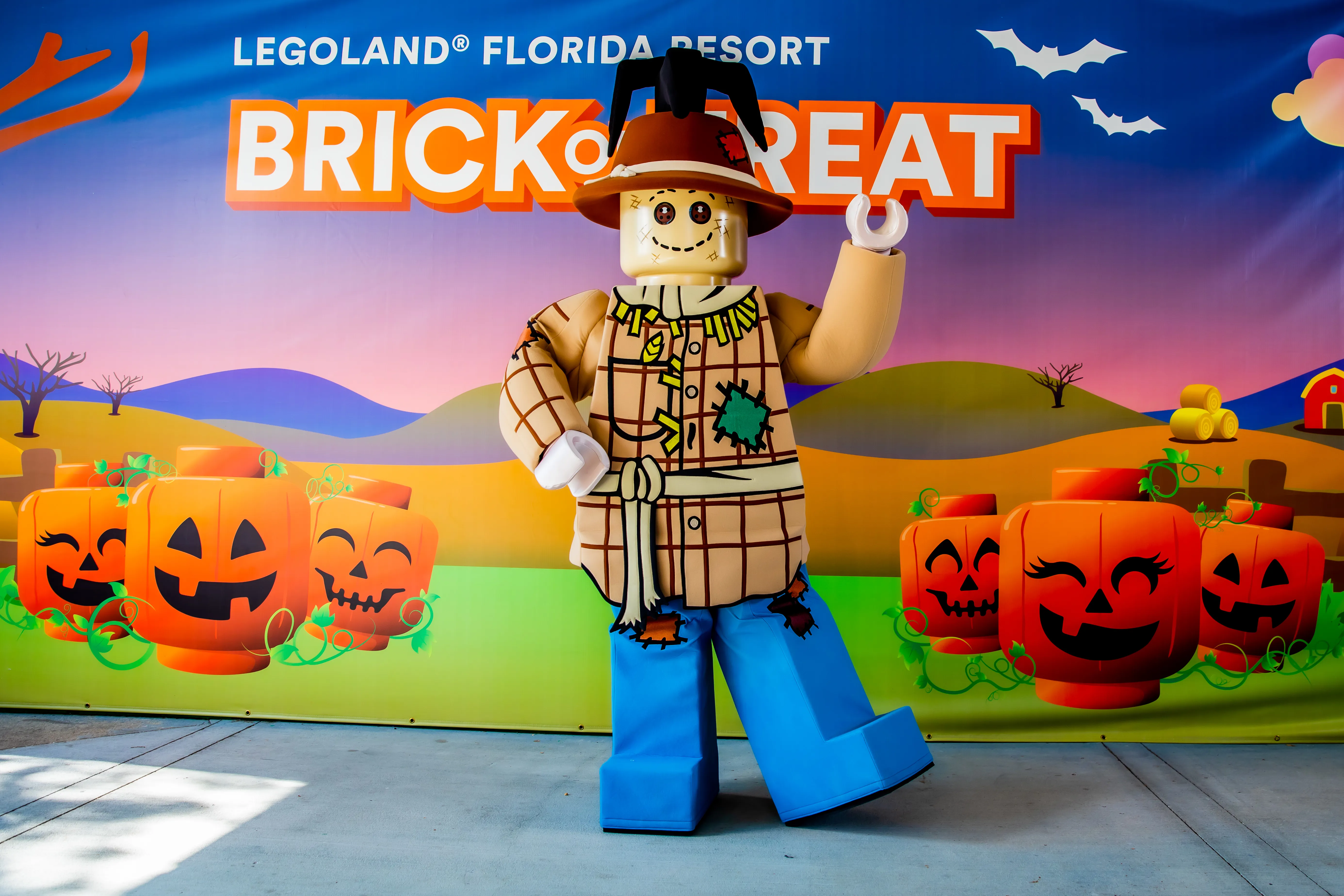 The walkaround LEGO Scarecrow character posed in front of a Brick or Treat photo backdrop, wearing a plaid shirt with a green patch, a brown hat with a black crow on the crown, and blue legs, one hand raised. The backdrop shows rows of orange jack o lantern minifigure heads, rolling hills, a small red barn and the words Legoland Florida Resort Brick or Treat