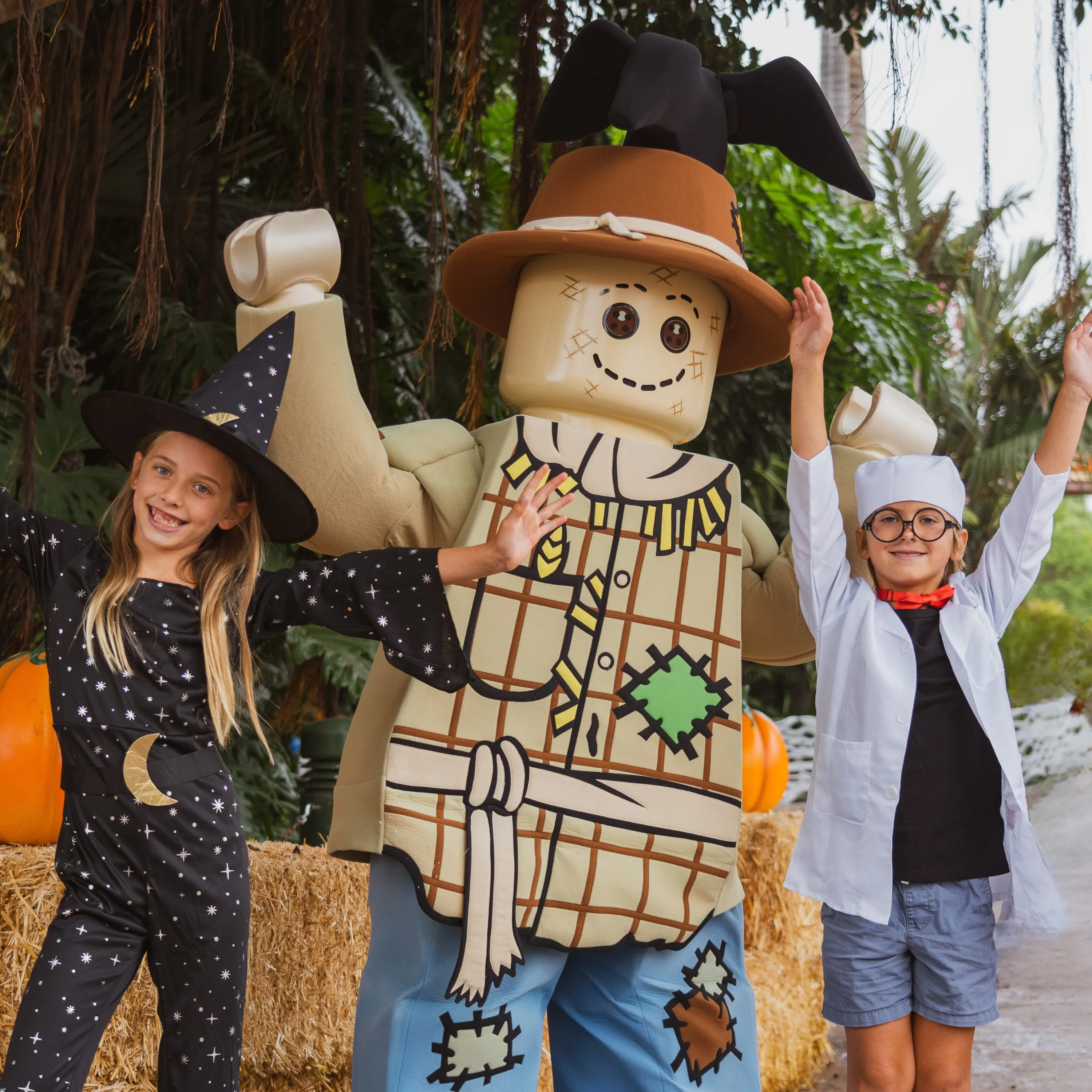The walkaround LEGO Scarecrow character stands between two children in Halloween costumes in front of hay bales and pumpkins, one girl in a black witch hat and star patterned outfit and one boy in a white lab coat and round glasses, both with arms raised, and a third child's raised arm just visible at the left edge. A black LEGO crow sits on the crown of the scarecrow's brown hat