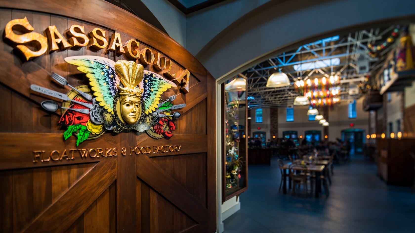 A carved wooden entrance sign for Sassagoula Floatworks and Food Factory: a gilded Mardi Gras mask with a winged crest and cutlery, and the lit dining room of the food court visible through the archway to the right
