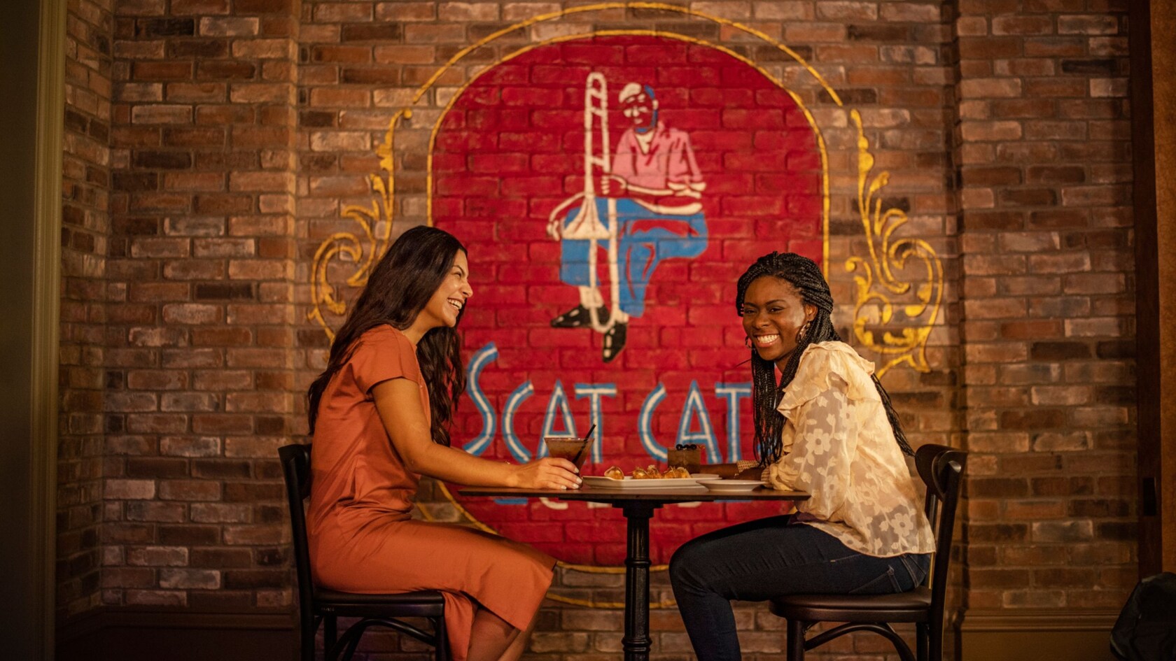 Two guests seated at a small bistro table with drinks and a plate of appetizers, in front of a painted red and gold mural of a seated jazz musician in a flat cap playing a trombone on an exposed brick wall