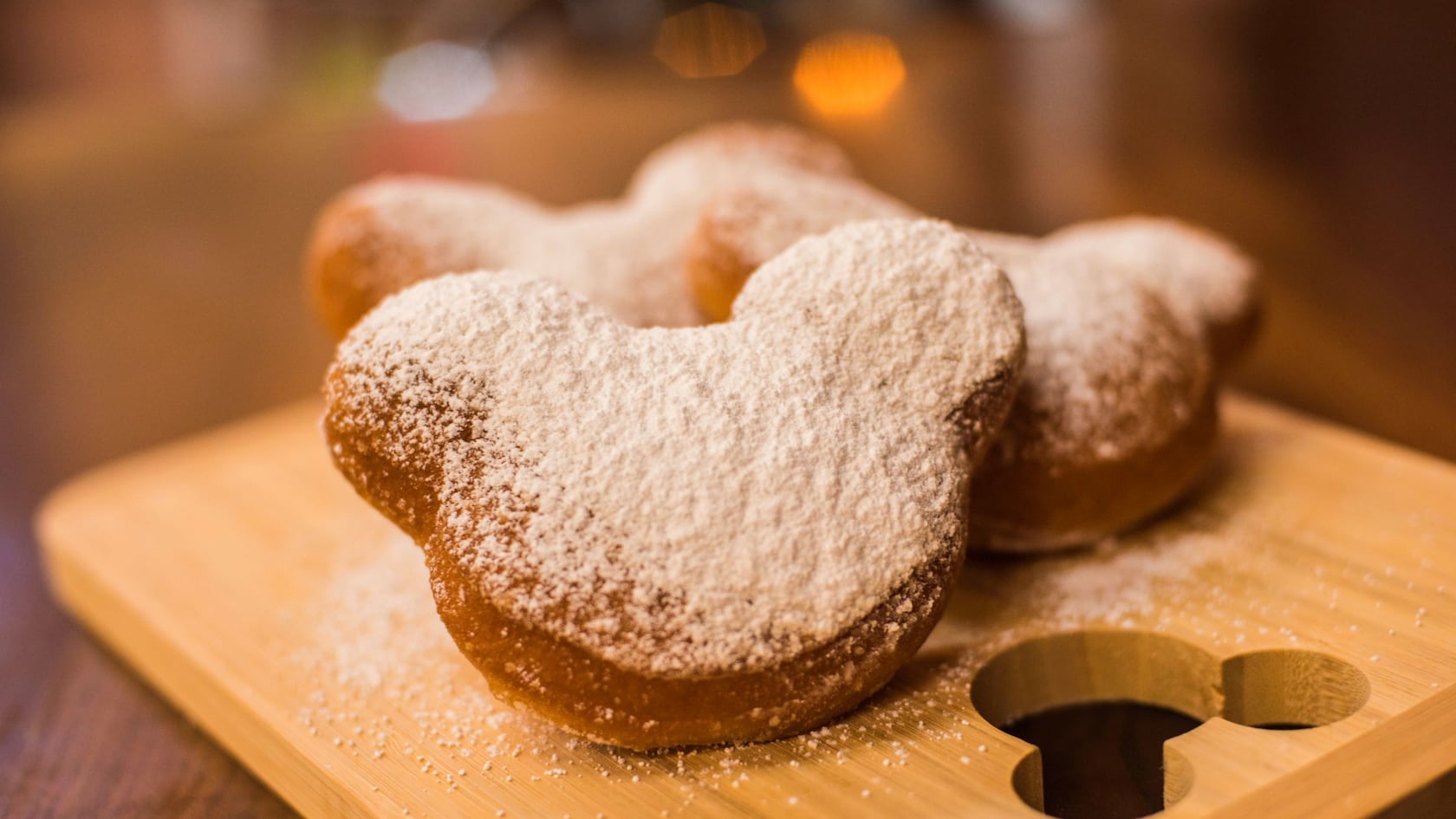 Three Mickey-shaped beignets heavily dusted with powdered sugar on a bamboo serving board with a Mickey-shaped cutout holding dipping sauce