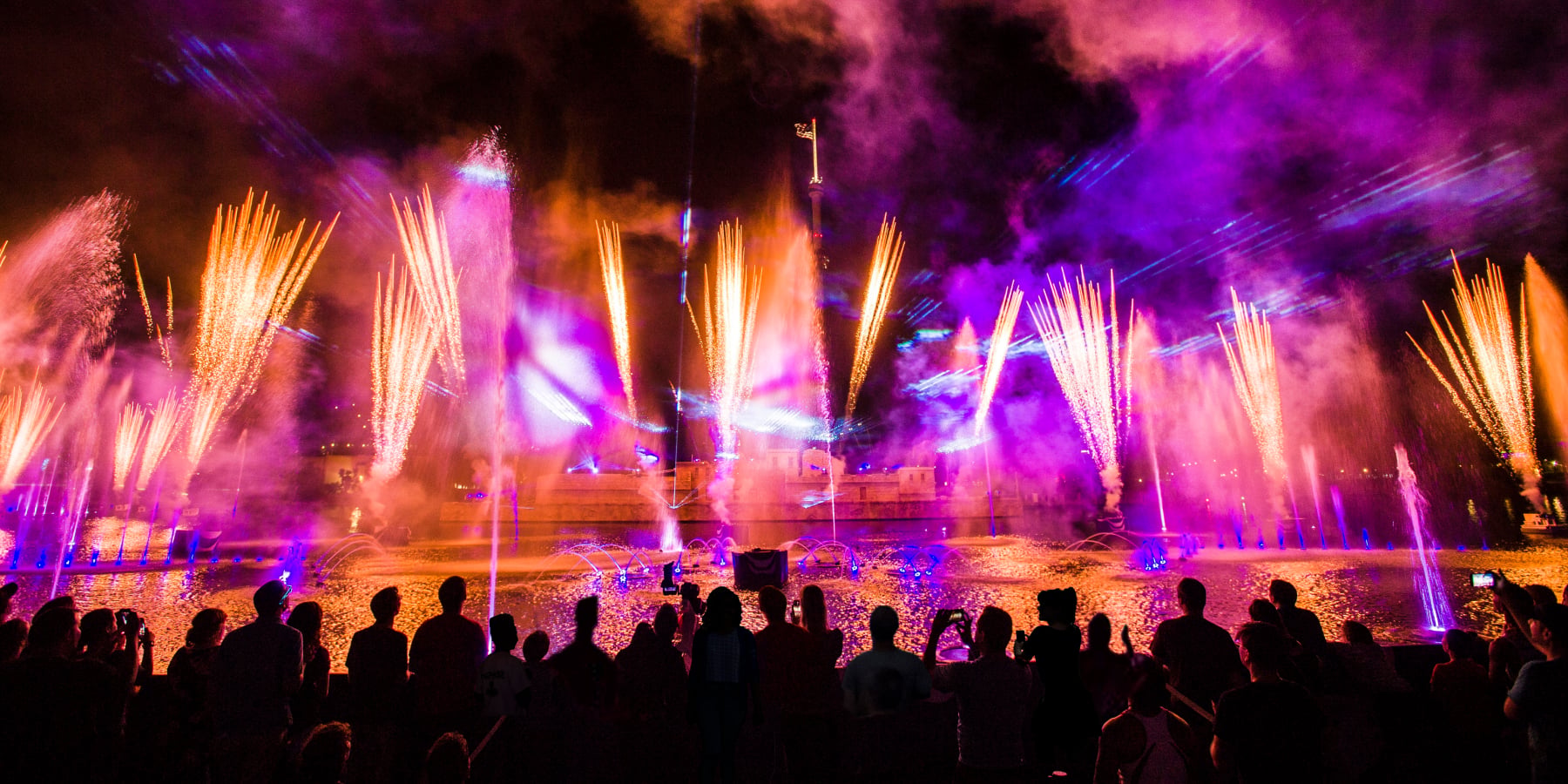 A crowd of silhouetted guests at a waterside rail watching a nighttime show, with tall gold firework fountains, lit blue water jets on the lagoon, blue and purple laser beams and magenta lit smoke above a low terraced stone structure on the far shore
