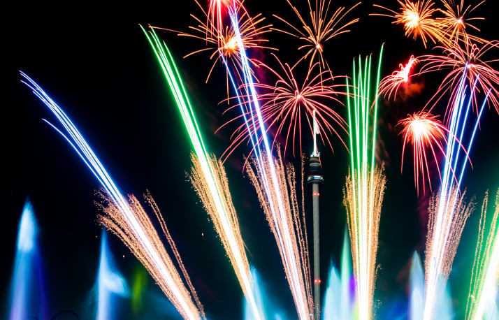 Fireworks in green, blue, gold and magenta bursting around the silhouette of SeaWorld's Sky Tower against a black night sky, with gold comet trails rising from below