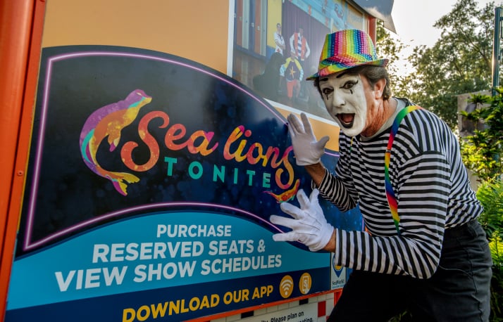 A performer in mime makeup, a rainbow sequined fedora, striped shirt and white gloves posing with hands raised beside a colorful Sea Lions Tonite sign reading Purchase Reserved Seats and View Show Schedules