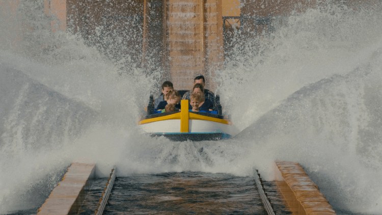 A head on view from water level of a white and yellow boat of riders at the bottom of the drop, twin sheets of water fanning up on both sides of the hull, with the brown drop track rising behind them under flat grey light