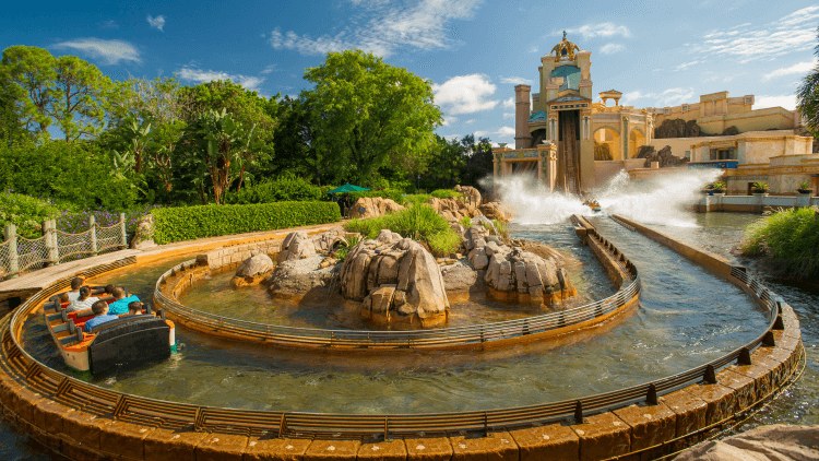 A wide daylight view of Journey to Atlantis, with the ride's water channel curving around a low rockwork island in the foreground, a boat of riders gliding along it at left, tropical plantings and trees behind them, and the Atlantis tower and a tall white splash plume in the background under a partly cloudy sky